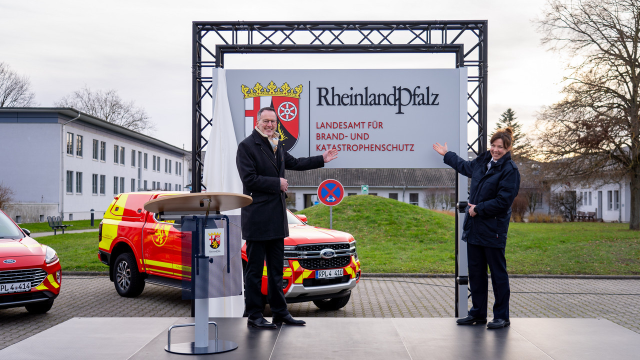 Innenminister Michael Ebling und Landesbrand- und Katastrophenschutzinspekteurin Kirstin Eisenhauer mit dem Schild der neuen Behörde. Im Hintergrund stehen landeseigene Einsatzfahrzeuge im neu gestalteten Design.