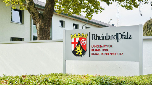 Das Schild des Landesamtes für Brand- und Katastrophenschutz steht vor einer Mauer mit einem Baum und dem Behördengebäude dahinter.